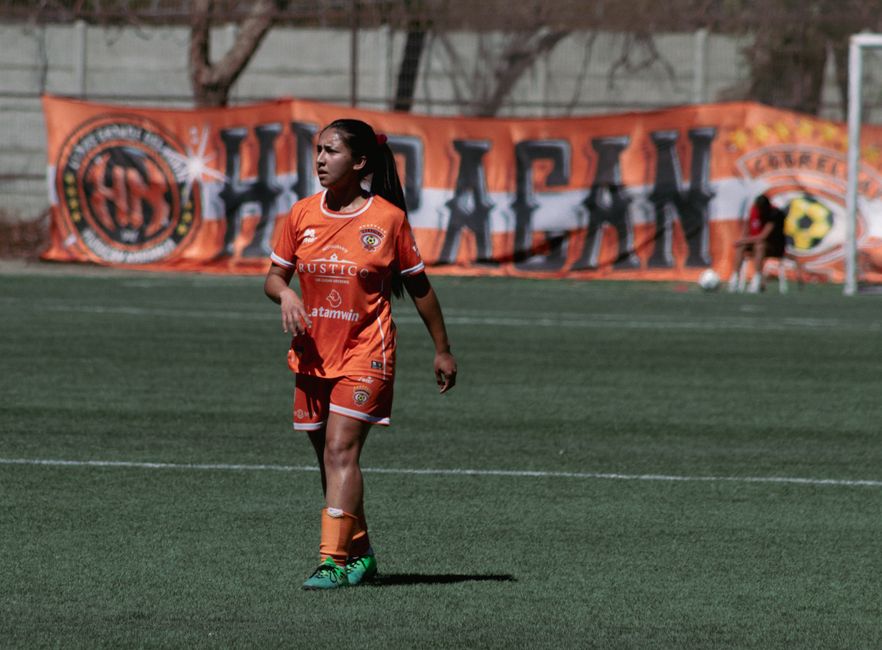 Female soccer player in orange uniform on outdoor field with banner background.