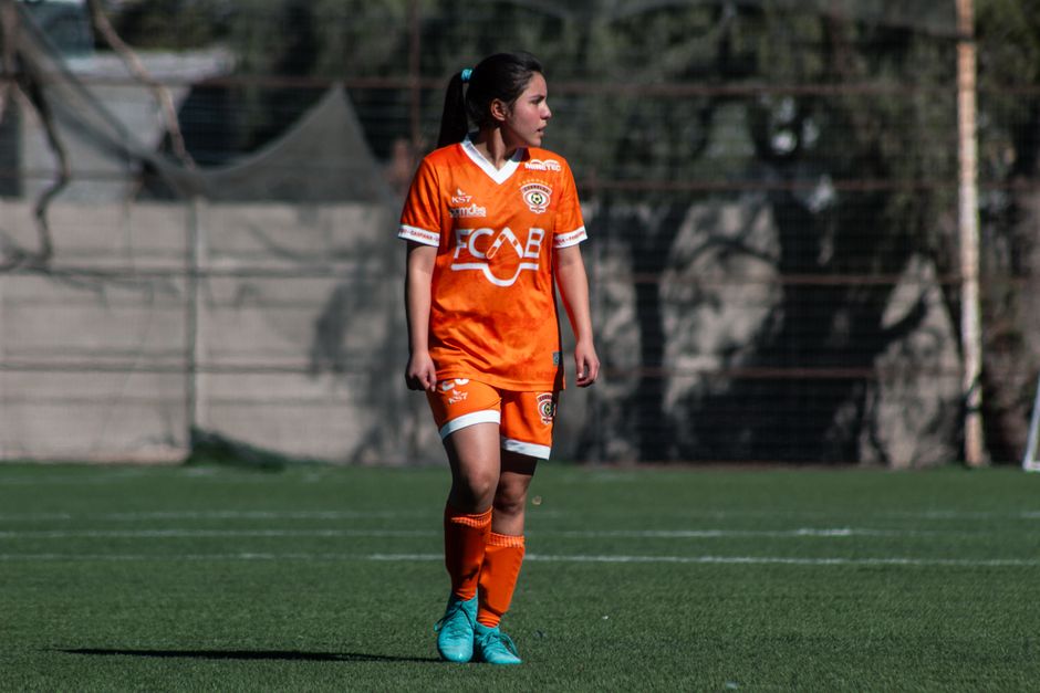 A female soccer player in orange uniform during a match on an outdoor field in Chile.