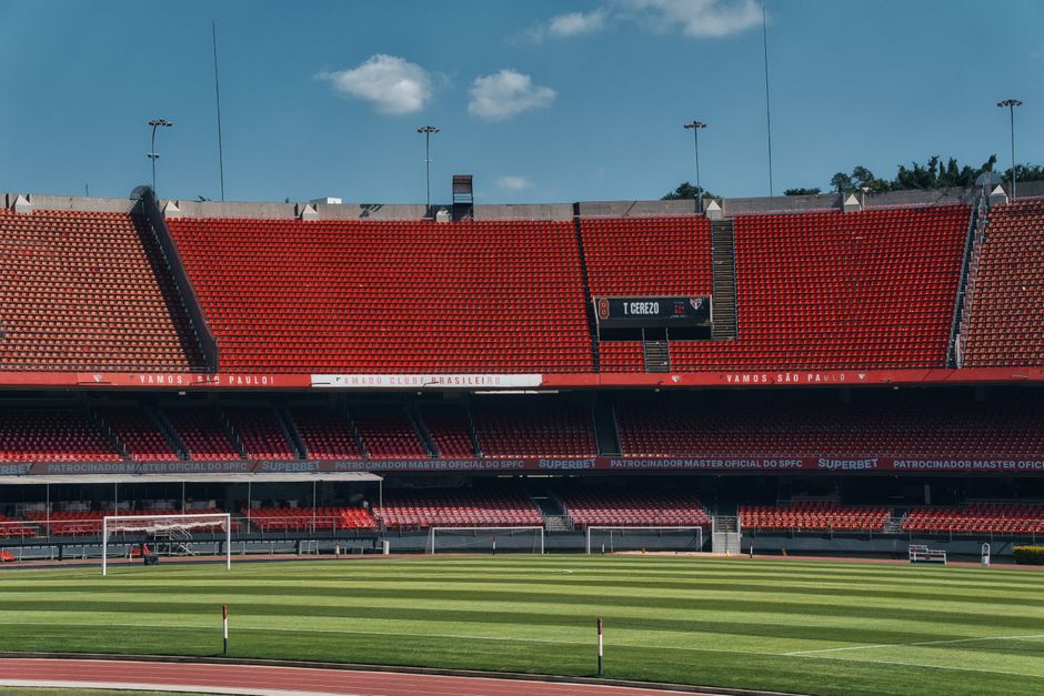 Wide view of an empty soccer stadium with red seating and green field under clear sky.