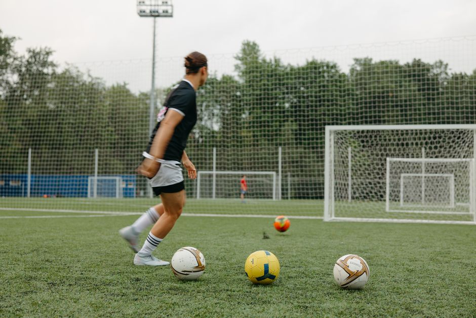 A woman practicing soccer on an outdoor field with multiple soccer balls and goals.
