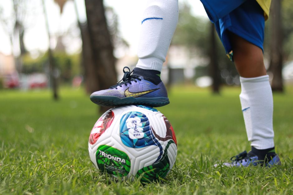 A young soccer player wearing white socks stands on a colorful soccer ball outdoors.